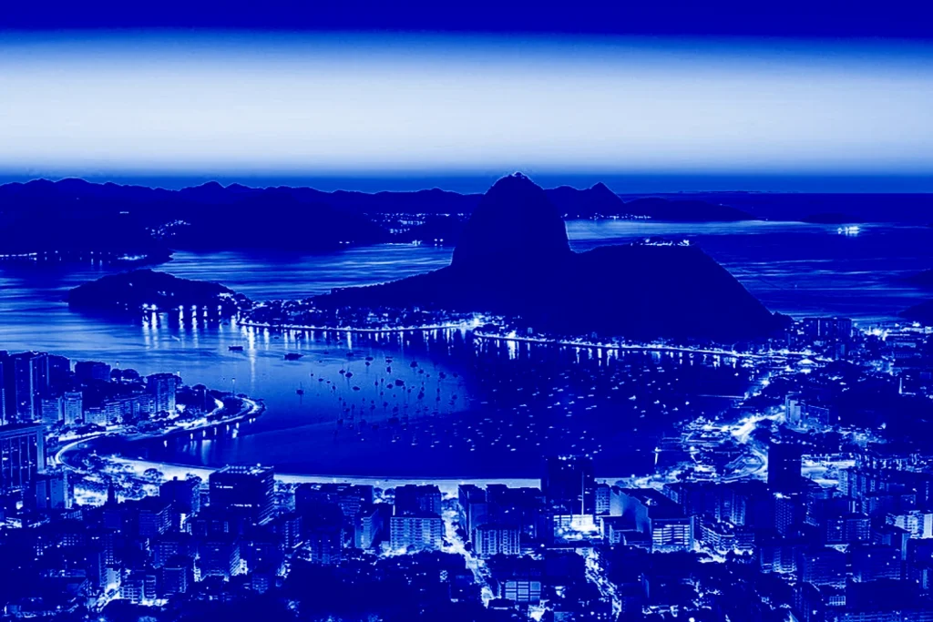 Vista aérea noturna em tons de azul do Rio de Janeiro, destacando a Enseada de Botafogo e o Pão de Açúcar iluminado.